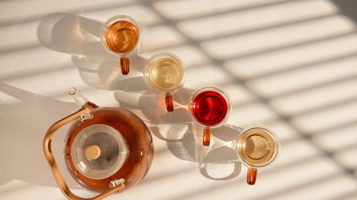 A glass teapot of brewed tea beside four glass cups, one of each blend, from pale gold to berry red, in striped window light