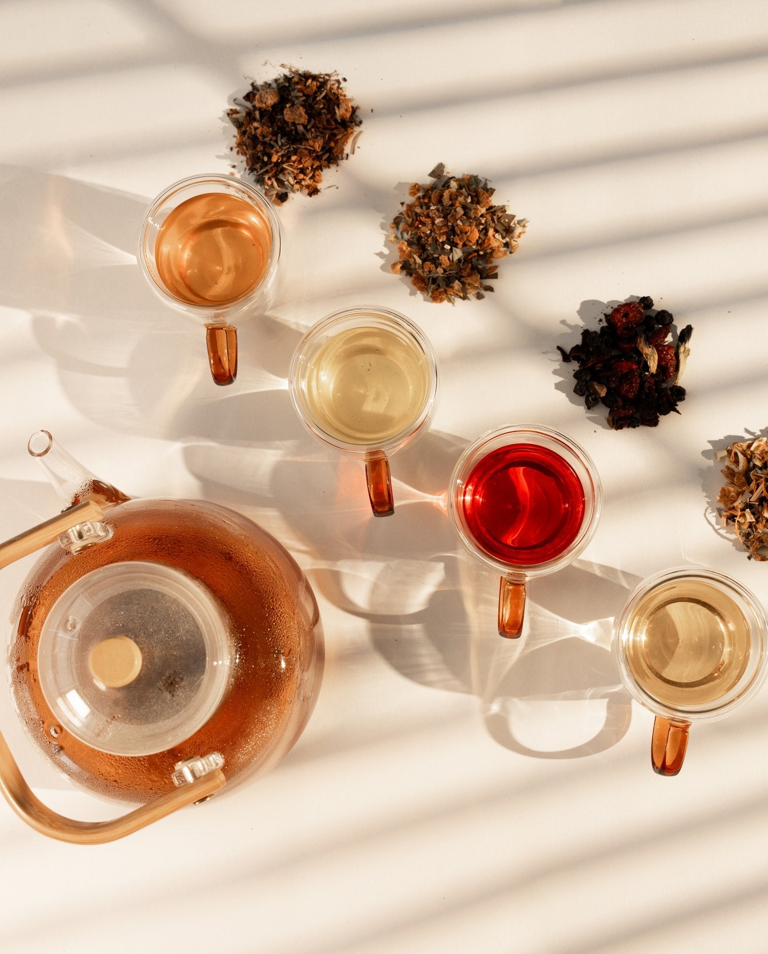 Overhead, a glass teapot beside four cups of brewed tea and a pile of each loose blend