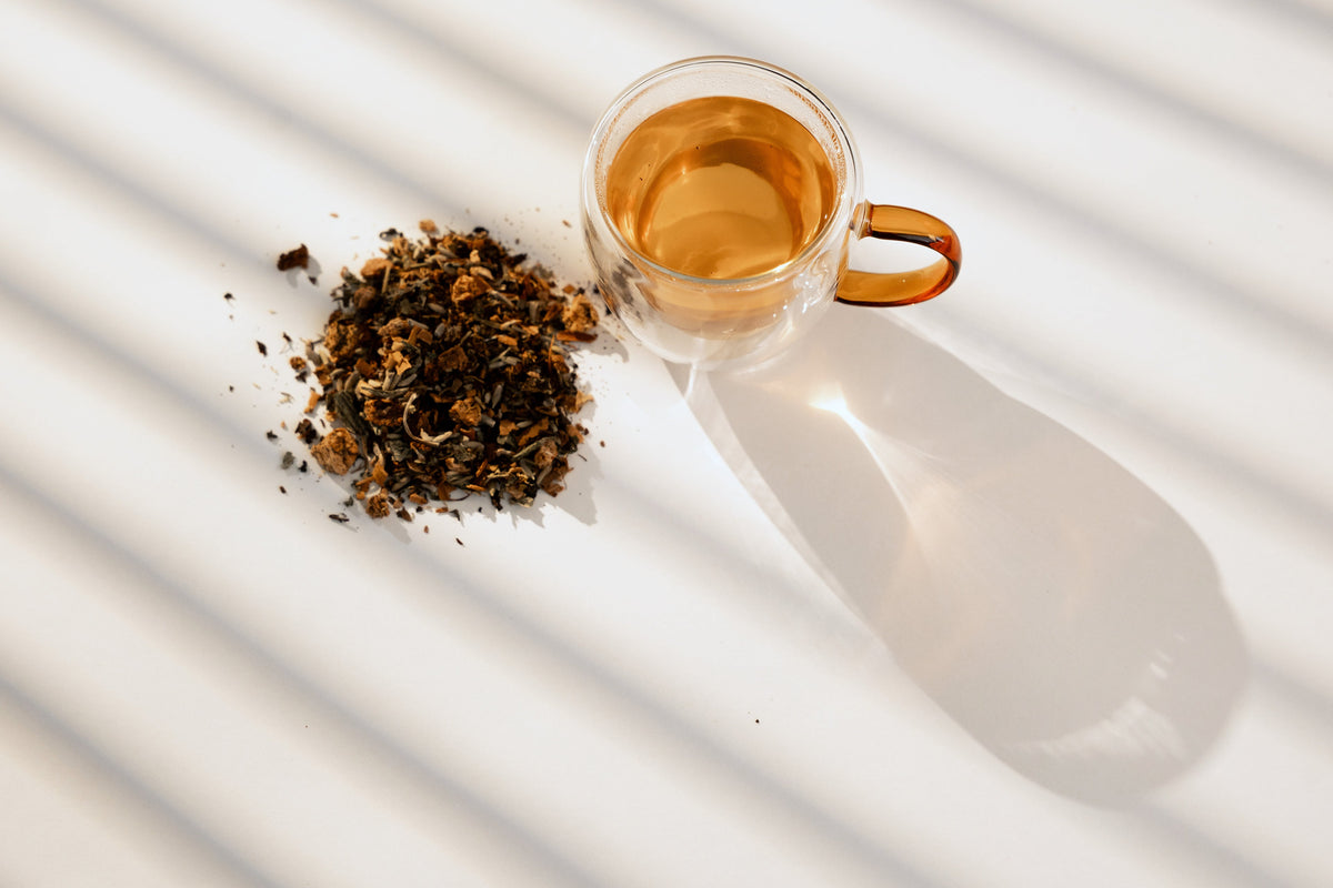 A glass cup of brewed Dusk beside a pile of the dry blend on a white surface under striped sunlight