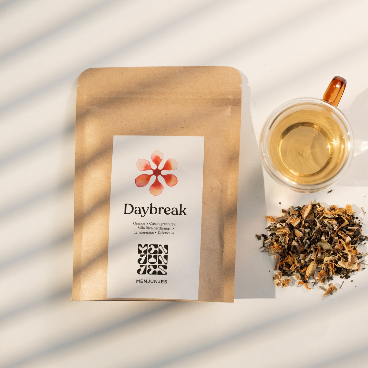 The Daybreak pouch beside a glass cup of the brewed tea and a pile of the loose blend, in striped window light