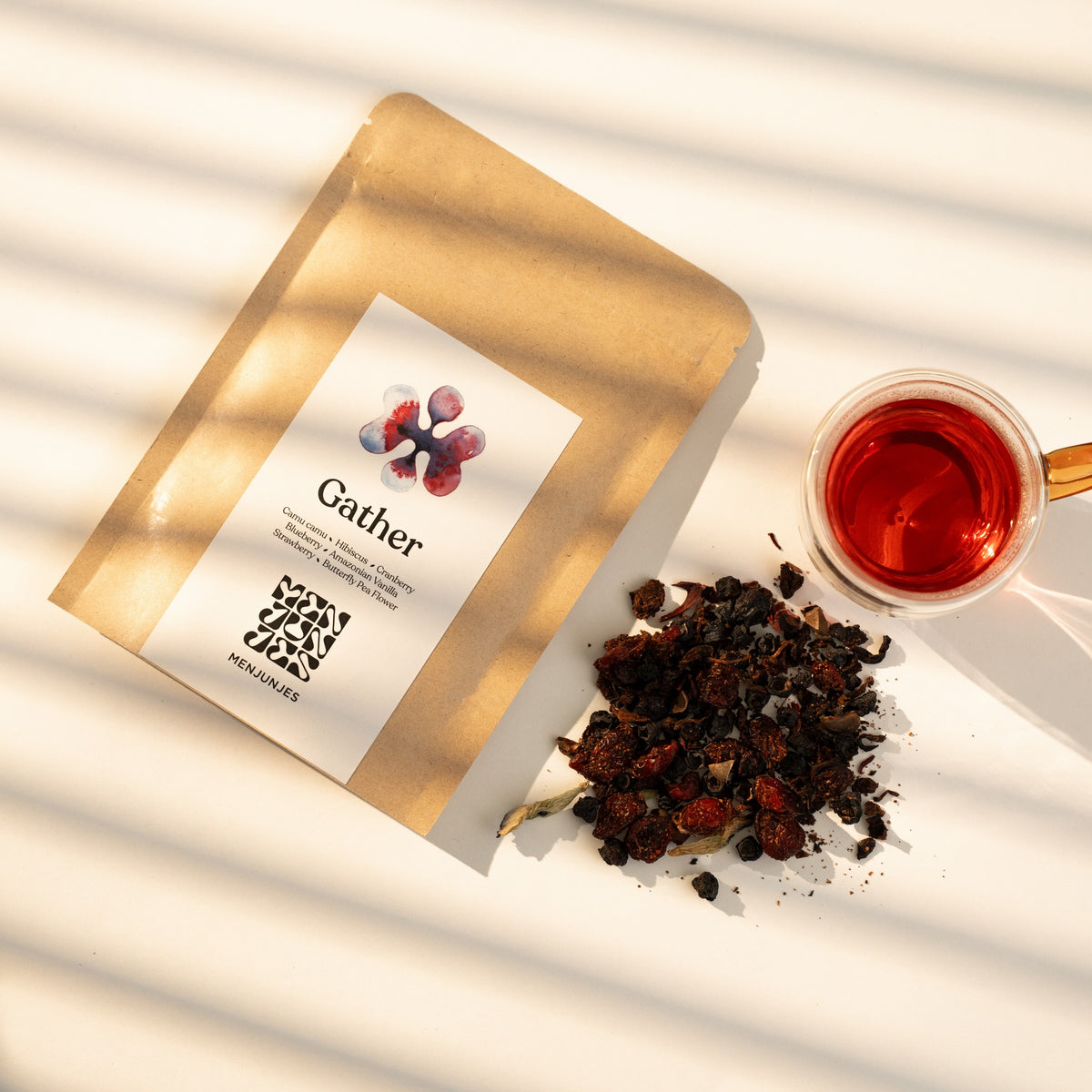 The Gather pouch beside a glass cup of the brewed tea, deep berry red, and a pile of the loose blend