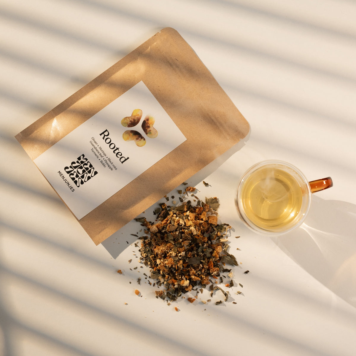 The Rooted pouch beside a glass cup of the brewed tea and a pile of the loose blend, in striped window light